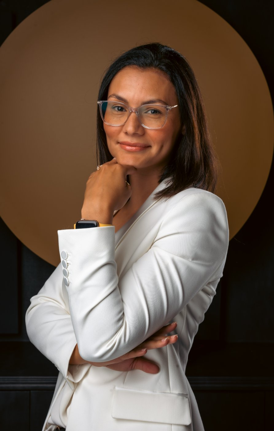 Lynn in a white blazer and clear-framed glasses, arms folded in front of a warm amber circle, with a calm and assured expression.
