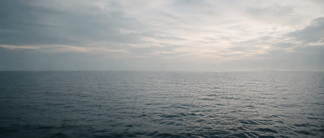 A calm, open sea meeting a soft, cloud-veiled horizon in muted dawn light.
